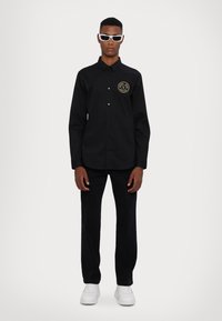 Camicia nera a maniche lunghe con bottoni e un'emblema dorato rotondo, abbinata a pantaloni neri e sneakers bianche. Modello con occhiali da sole bianchi.