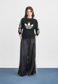 Svart strikkegenser med hvit tre-stripes design og stor Adidas-logo, kombinert med sorte sateng brede bukser med sidestriper.