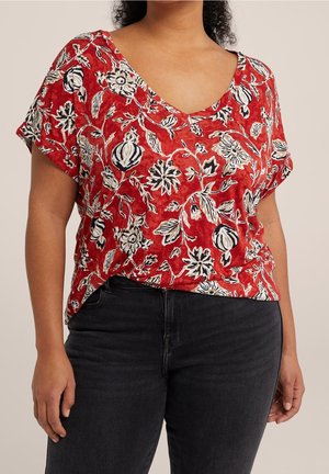 Vrouw draagt een rood V-hals top met witte en zwarte bloemmotieven, gecombineerd met zwarte jeans, tegen een effen achtergrond.
