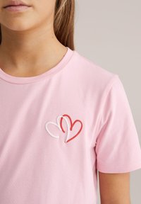 T-shirt in cotone rosa con ricamo di un cuore rosso e bianco sull'area sinistra del petto, maniche corte e scollo a girocollo.