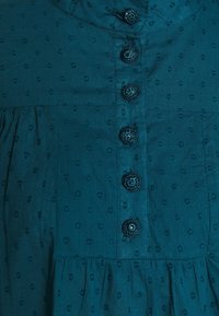 Blåt stof med små teksturerede prikker, der har seks dekorative runde hækleknapper ned langs midten og sammentrækningsting under knapperne.