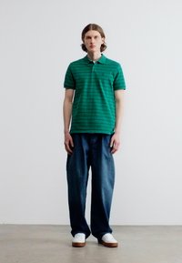 Jeune homme portant un polo rayé vert, un jean bleu foncé ample et des baskets blanches, debout contre un mur blanc uni.