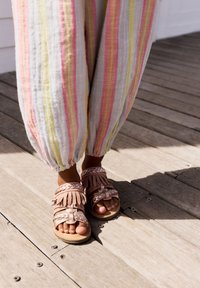 Sandali rosa chiaro con dettagli in frange e accenti intrecciati, abbinati a pantaloni leggeri a righe nei toni pastello, su una superficie di legno.