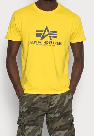 Gul bomulds T-shirt med blåt logo "Alpha Industries" og stjerne design, sammen med grøn camouflage cargo shorts med lommer og mønster.