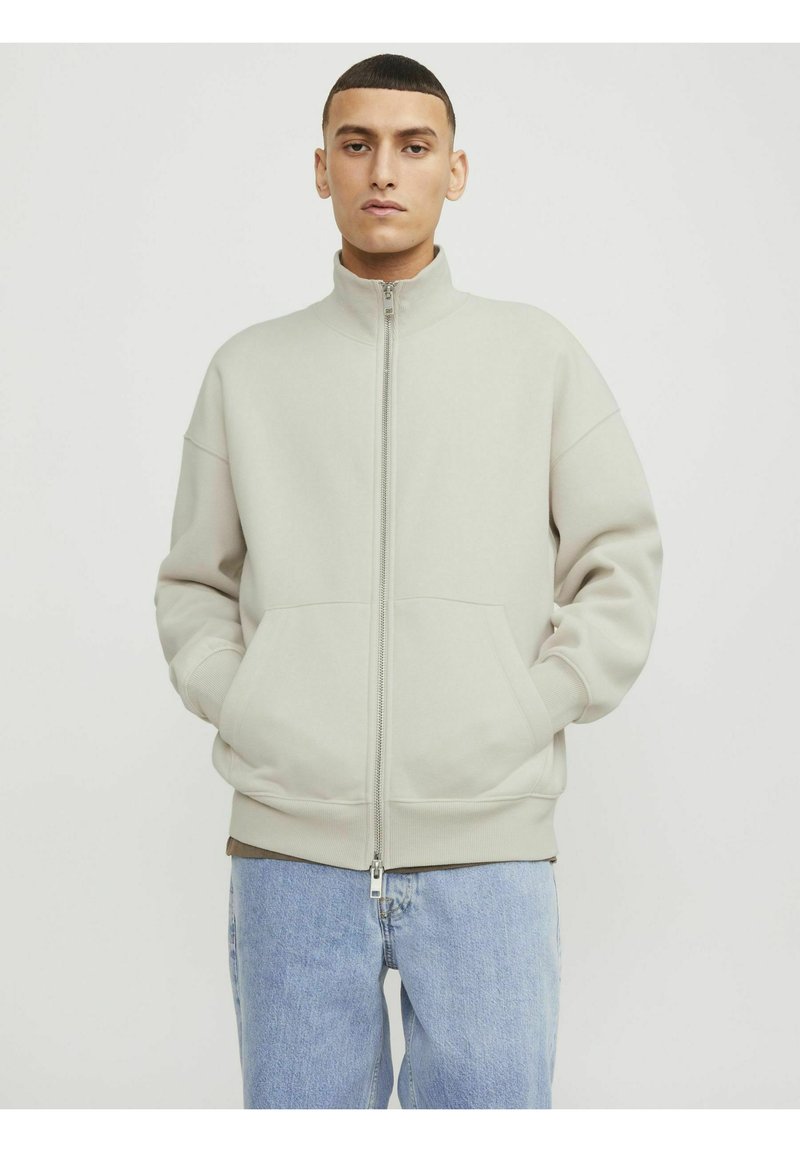 Lys grå zip-up jakke med høj krave, drop skuldre, to forlommer og ribbede manchetter. Båret sammen med lyseblå jeans.