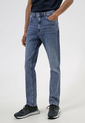 Mann trägt schmal geschnittene, leicht abgenutzte blaue Jeans und schwarze Sportschuhe, steht mit einer Hand in der Tasche.