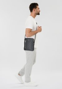 Bolso cruzado gris con un diseño estampado, cierre de cremallera y correa ajustable, llevado al lado por un hombre en ropa casual. Presenta un logo triangular.