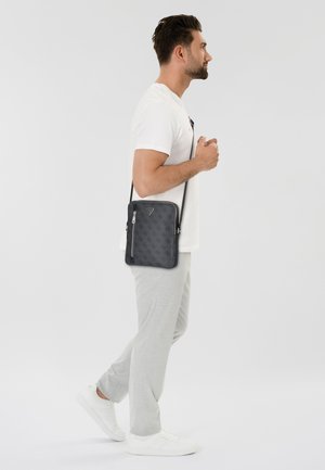 Sac bandoulière à motif gris, avec fermeture éclair et sangle réglable, porté sur le côté par un homme en tenue décontractée. Présente un logo triangulaire.