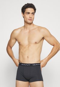 Boxeri din bumbac de culoare gri închis, cu un tiv elastic pe care este inscripționat numele brandului „GANT” în alb. Modelul pozează cu o expresie neutră.