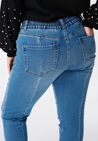 Jeans en denim bleu avec une taille élastique, deux poches arrière et un design slim fit. Le tissu a une surface lisse et légèrement texturée.