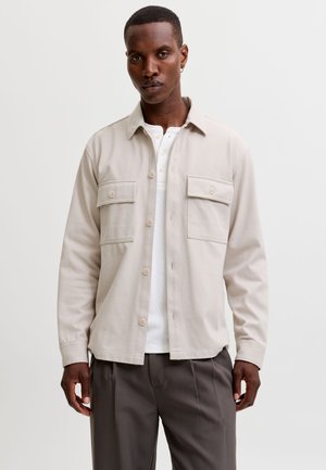 Homme portant une veste-chemise beige à boutons sur une chemise henley blanche et un pantalon foncé, debout devant un fond clair uni.