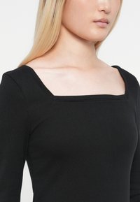 Crna majica dugih rukava s četvrtastim izrezom, izrađena od glatkog, rastezljivog materijala. Dizajn ima prilagođen siluetu i suptilne šavove.