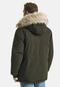 Parka vert olive avec une capuche doublée en fourrure, dotée d'une fermeture éclair, de poches latérales et de poignets côtelés. Le tissu texturé offre chaleur.