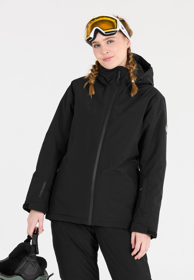 Schwarze wasserdichte Jacke mit hohem Kragen, Reißverschluss vorne und verstellbarem Kapuze. Trägt Schneeg goggles und hält einen Skistiefel in einer Hand.