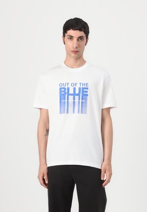 Uomo con capelli corti e scuri che indossa una maglietta bianca con scritta blu "OUT OF THE BLUE" e pantaloni neri, in piedi davanti a uno sfondo bianco uniforme.