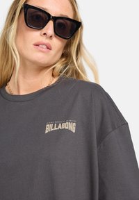 Mørkegrå T-shirt med rund hals, der har et guld Billabong-logo på brystet. Stoffet virker blødt og har en afslappet pasform.