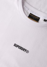 Camiseta de algodón blanca con cuello redondo y mangas cortas. Logo bordado en negro "SUPERDRY" en la parte frontal. Textura suave.