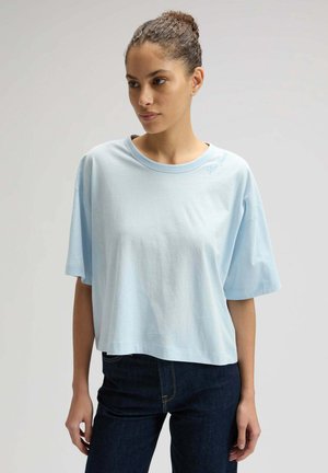 Frau mit lockigem Haar in einem locker sitzenden hellblauen T-Shirt und dunklen Jeans, die vor einem schlichten Hintergrund steht und leicht nach links schaut.