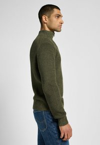 Oliivinvihreä ribbiknitti neule, jossa on korkea kaulus ja tiukka siluetti, yhdistettynä sinisiin farkkuihin. Huomattava tekstuurinen yksityiskohta hihoissa.