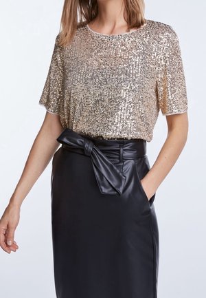 Mujer con una camiseta de lentejuelas doradas de manga corta metida en una falda de cuero negra de cintura alta con cinturón atado.