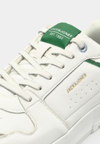 Bijele kožne tenisice s zelenim naglaskom na jeziku, s perforiranim detaljima, bijelim vezicama i zlatnim logotipom sa strane.