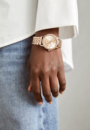 Montre en or rose avec un bracelet en métal texturé, cadran rond et trois sous-cadrans, accentuée par une lunette en cristal. Portée sur une main contre du denim.