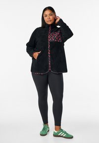 Schwarze Fleecejacke mit roten Polka-Dot-Akzenten, durchgehendem Reißverschluss, Seitentaschen und abgerundetem Saum, kombiniert mit schwarzen Leggings und grünen Sneakers.