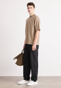 Tan short-sleeve top with a crew neck, paired with dark denim pants and white sneakers. Holding a brown bag. Minimalist design, casual look.