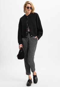 Schwarze, kurze Strickjacke mit Rundhalsausschnitt und Knöpfen, graue strukturierte Hose, schwarze Slipper und eine kleine schwarze, flauschige Handtasche.
