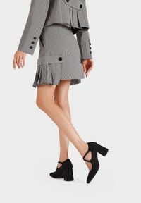 Chaussures à talons hauts en suede noir avec un bout rond, une sangle structurée à la cheville et un talon bloc épais ; matériau texturé ; design minimaliste.