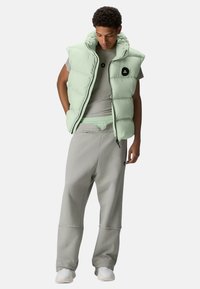 Hellgrüne gesteppte Weste mit hohem Kragen, getragen über einem grauen, eng anliegenden T-Shirt. Graue Jogginghosen mit elastischem Bund und Taschen.