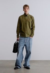 Πράσινο πουκάμισο με κουμπιά και τσέπη στο στήθος, ανοιχτό μπλε τζιν με φαρδιά πόδια, και μαύρα δερμάτινα παπούτσια, κρατώντας μια μαύρη σκληρή τσάντα χειρός.