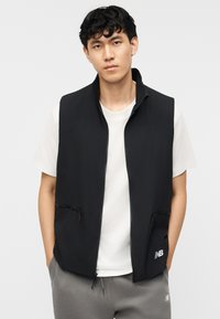 Νέος άνδρας που φοράει μαύρο γιλέκο με φερμουάρ πάνω από λευκό t-shirt και γκρι φόρμα, στέκεται με τα χέρια στις τσέπες μπροστά σε απλό φόντο.