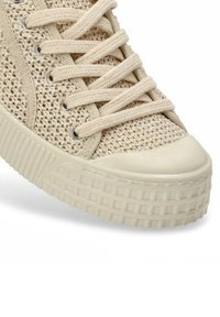 Zapatilla beige con parte superior de textil tejido, puntera redonda y suela gruesa y texturizada. Presenta cordones planos y ojales plateados.