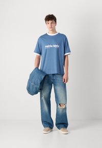 Blue short-sleeve T-shirt with white ribbed accents, featuring "calvin klein" logo printed on the front. Worn with distressed blue jeans.