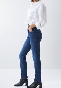 Mujer vistiendo jeans ajustados azules, botines negros de tacón y una camisa blanca de botones con las manguitas remangadas.