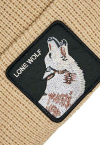 Beanie in maglina beige caratterizzato da un patch quadrato nero con una grafica di un lupo e il testo "LONE WOLF" sopra di esso. Texture morbida, design semplice.