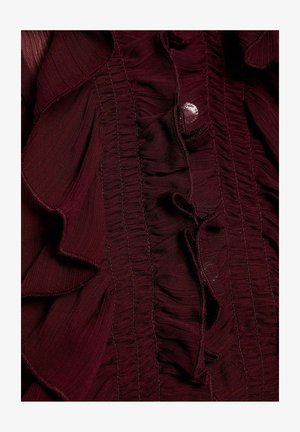 Blusa bordeaux con accenti arricciati, tessuto texture, dettagli increspati e chiusura con bottoni. Materiale liscio e leggero.