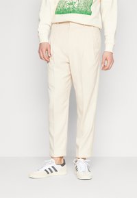 Jasbeżowe spodnie o dopasowanym kroju z zakładkami, zestawione z białymi trampkami z szarymi paskami. Gładka faktura.