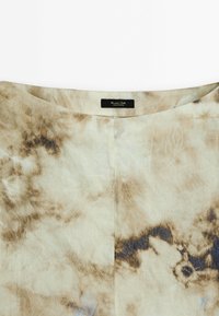 Tissu beige clair avec des motifs tie-dye marron et bleu, doté d'un col rond et d'une couture centrale à l'avant. Étiquette visible.