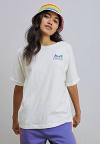 Witte oversized T-shirt met opgerolde mouwen, met een blauwe palmboomafdruk. Model draagt een meerkleurige emmerhoed en paarse broek.