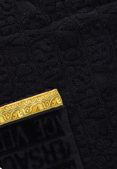 Tissu noir texturé avec des lettres en relief et un bord plié présentant une bordure décorative jaune avec des motifs ornés.