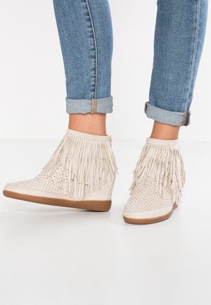 Bottes compensées beiges pour cheville avec détails à franges, design perforé, portées avec un jean bleu retroussé sur un fond de sol blanc.