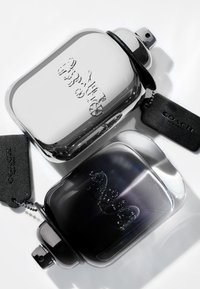 Due bottiglie di profumo rettangolari, una argentata e l'altra nera, presentano design in rilievo, tappi neri e etichette in pelle con dettagli "COACH".