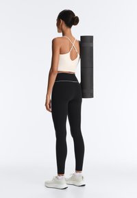 Kobieta w czarnych legginsach, białym krótkim topie i trampkach, niosąca zwiniętą czarną matę do jogi na plecach, zwrócona tyłem.