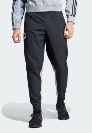 Mann trägt schwarze Jogginghose mit rosa und weißen Seitenteilen und schwarzen Sneakers vor einfarbigem Hintergrund.