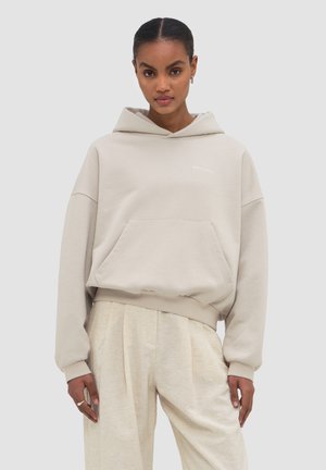 Frau trägt hellbeigen oversized Hoodie mit Fronttasche und passenden lockeren Hosen, steht vor einfarbigem Hintergrund.