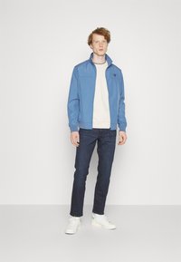 Giacca zip-up azzurra chiara con colletto alto, sotto una maglietta bianca a girocollo, jeans scuri e sneakers bianche con dettagli verde.