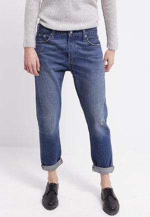 Person iført op-rullede blå jeans, sorte snørestøvler og en lysegrå langærmet sweater, stående mod en ensfarvet baggrund.