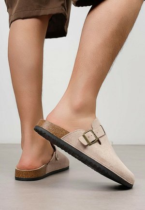 Personne portant des sabots beige à enfiler avec une boucle, une semelle intercalaire en liège marron et une semelle noire, debout sur un sol lisse avec les jambes visibles.